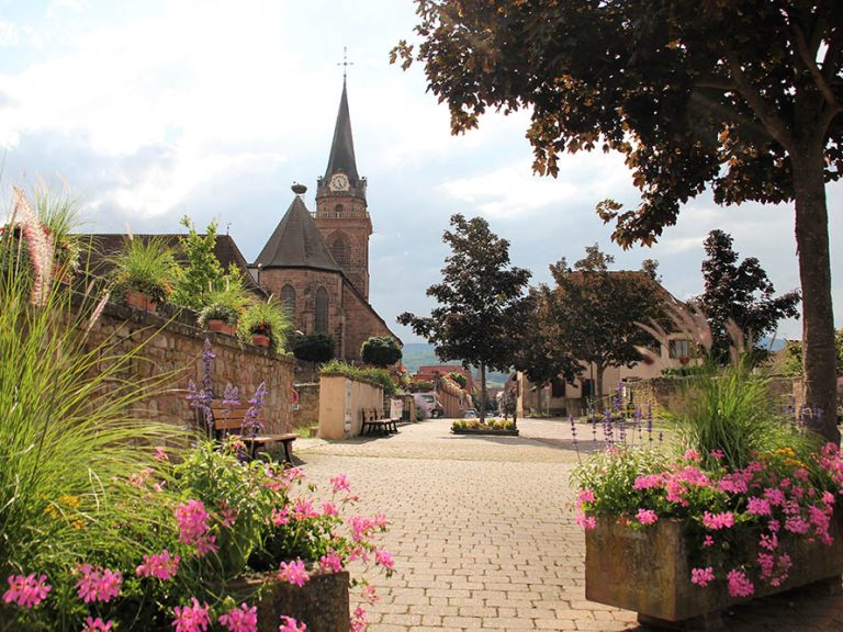 Place majoritairement minérale, avec des bacs à fleurs et quelques arbres avec en arrière plan l'église du village.